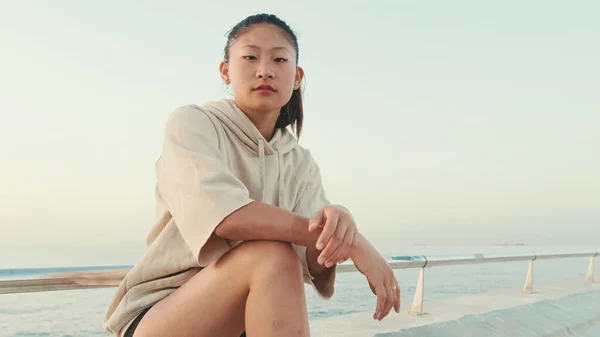 Asian girl wears sportswear sits on the promenade at morning time, on the background of the sea