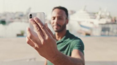 Genç adam arka planda yatlarla limanda otururken telefonda selfie çekiyor.