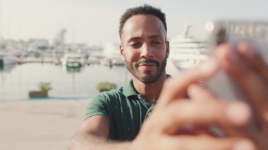Genç adam arka planda yatlarla limanda otururken telefonda selfie çekiyor.