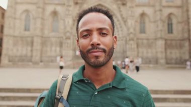 Omzunda sırt çantasıyla, kameraya bakıp eski şehrin meydanında dururken gülümseyen genç turistin yakın çekimi.