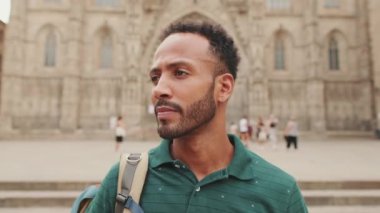 Omzunda sırt çantası olan genç turist, eski şehrin meydanında dikilirken kameraya bakıyor.