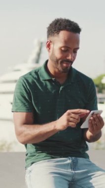 Genç gülümseyen adam arka planda yatlarla limanda otururken telefonu kullanıyor.