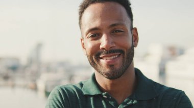 Yakın plan, genç adam arka planda yatlarla limanda oturuyor, kameraya bakıyor ve gülümsüyor.