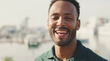 Yakın plan, genç adam arka planda yatlarla limanda oturuyor, kameraya bakıyor ve gülümsüyor.