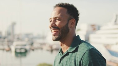 Genç gülen adam limanda arka planda yatlarla oturuyor.