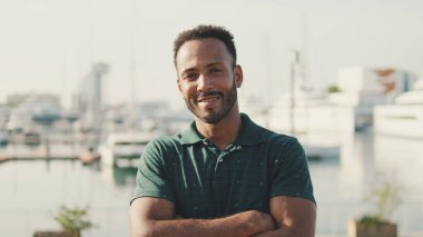 Arka planda yatlarla limanda oturan genç adam kameraya bakıyor ve gülümsüyor.