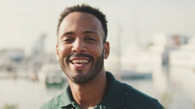 Yakın plan, genç adam arka planda yatlarla limanda oturuyor, kameraya bakıyor ve gülümsüyor.