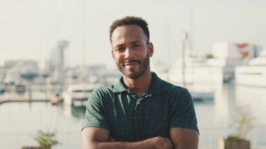 Arka planda yatlarla limanda oturan genç adam kameraya bakıyor ve gülümsüyor.