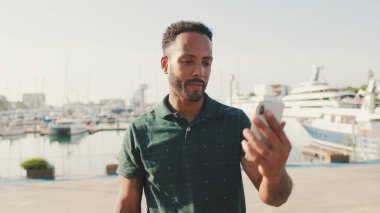 Limanda arka planda yatlarla otururken telefondan video görüşmesi yapan mutlu genç adam.