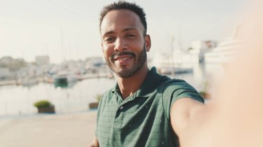 Limanda arka planda yatlarla otururken telefondan video görüşmesi yapan mutlu genç adam.