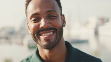 Yakın plan, genç adam arka planda yatlarla limanda oturuyor, kameraya bakıyor ve gülümsüyor.