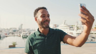 Limanda arka planda yatlarla otururken telefondan video görüşmesi yapan mutlu genç adam.