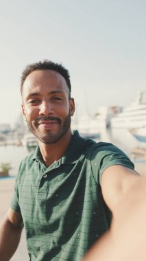 Limanda arka planda yatlarla otururken telefondan video görüşmesi yapan mutlu genç adam.