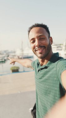 Limanda arka planda yatlarla otururken telefondan video görüşmesi yapan mutlu genç adam.