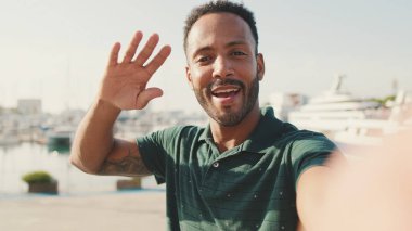 Limanda arka planda yatlarla otururken telefondan video görüşmesi yapan mutlu genç adam.