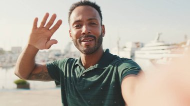 Limanda arka planda yatlarla otururken telefondan video görüşmesi yapan mutlu genç adam.