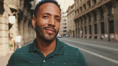 Sokakta dikilen mutlu genç adama yakın çekim. Adam kafasını çevirir ve kameraya gülümser.