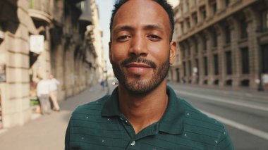 Sokakta dikilen mutlu genç adama yakın çekim. Adam kafasını çevirir ve kameraya gülümser.