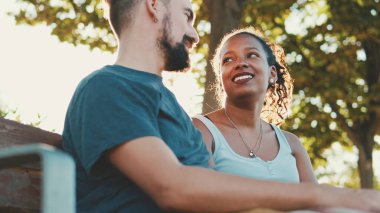 Interracial couple talking while sitting on bench. Backlight