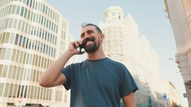 Close up, smiling young man with beard talking on the phone on modern buildings background