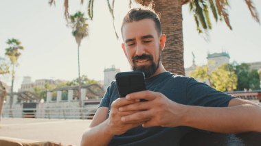 Young smiling man with beard sits on bench, uses cellphone