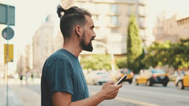 Young man with beard is using cellphone on the background of passing cars