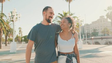 Happy interracial couple walking down the street talking and smiling. Man hugs the woman. 