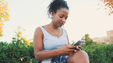 Laughing young mixed race woman sitting on park bench using cellphone