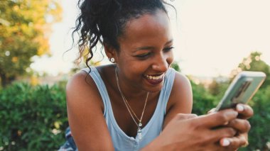 Laughing young mixed race woman sitting on park bench using cellphone