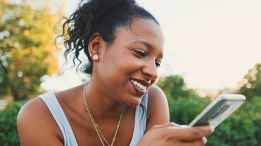 Smiling young mixed race woman sitting on park bench sending voice message