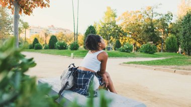 Young mixed race woman sitting on park bench, smiling, looking around