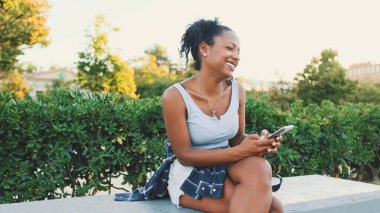 Laughing young mixed race woman sitting on park bench using cellphone