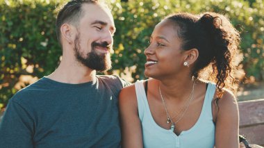 Interracial couple talking while sitting on bench. Backlight