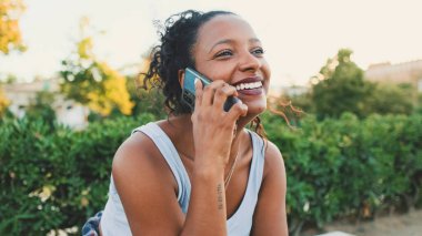 Laughing young mixed race woman sitting on park bench talking on cellphone