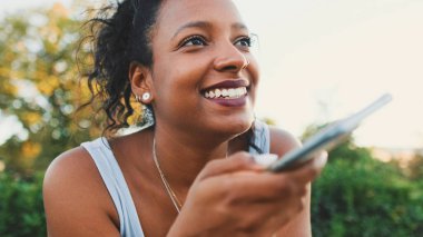 Smiling young mixed race woman sitting on park bench sending voice message