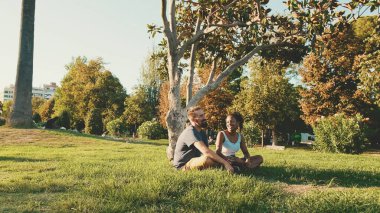 Happy smiling interracial couple talking while sitting on the grass in the park. Backlight