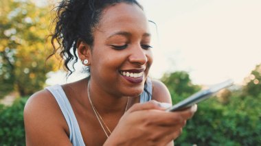 Smiling young mixed race woman sitting on park bench sending voice message