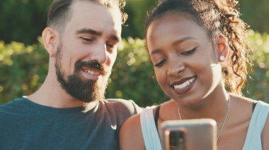Happy interracial couple talking while sitting on bench, using cellphone