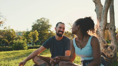 Happy smiling interracial couple talking while sitting on the grass in the park. Backlight