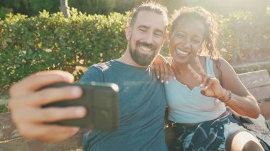 Happy smiling interracial couple taking selfie on phone while sitting on park bench. Backlight