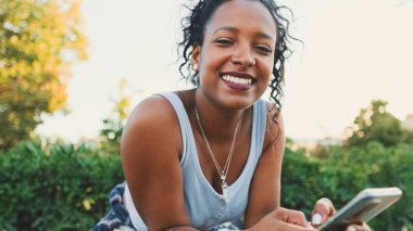 Close up, smiling young mixed race woman sitting on park bench using cellphone. Raises head looking at camera