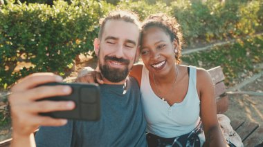 Happy smiling interracial couple making video call on cellphone while sitting on bench