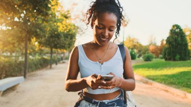 Laughing young mixed race woman walking along path in the park, using cellphone