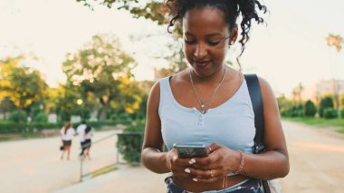 Laughing young mixed race woman walking along path in the park, using cellphone