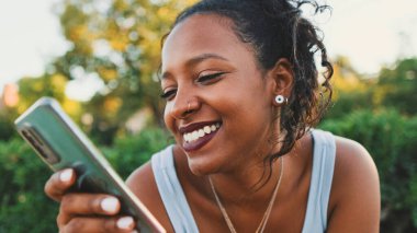 Smiling young mixed race woman sitting on park bench sending voice message