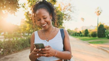Laughing young mixed race woman walking along path in the park, using cellphone