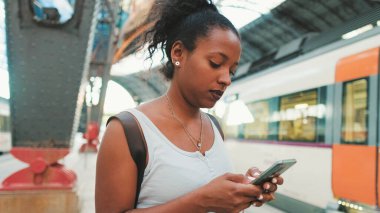 Close up, young mixed race woman stands at the railway station on the background of trains, uses smartphone