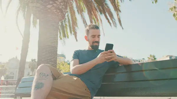 Young Smiling Man Beard Sits Bench Uses Cellphone — Zdjęcie stockowe