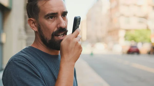 Close-up young man with beard uses cellphone while standing on crosswalk, sends voice message