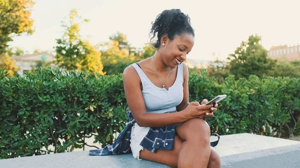 Laughing young mixed race woman sitting on park bench using cellphone